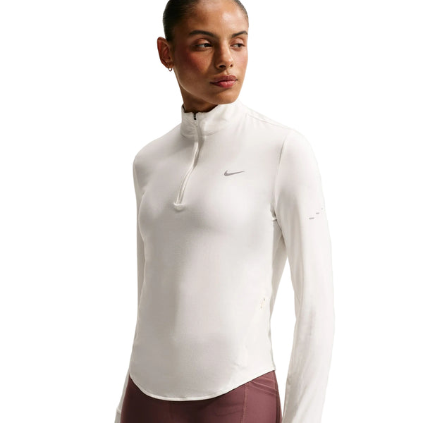 En person iført en hvid Nike Swift UV Half Zip-løbetrøje til kvinder i farven Sail/Reflective Silver, der er fremstillet af svedtransporterende stof og har en kvart lynlås, kombineret med mauve leggings, står med ansigtet let vendt til siden foran en ensfarvet hvid baggrund.