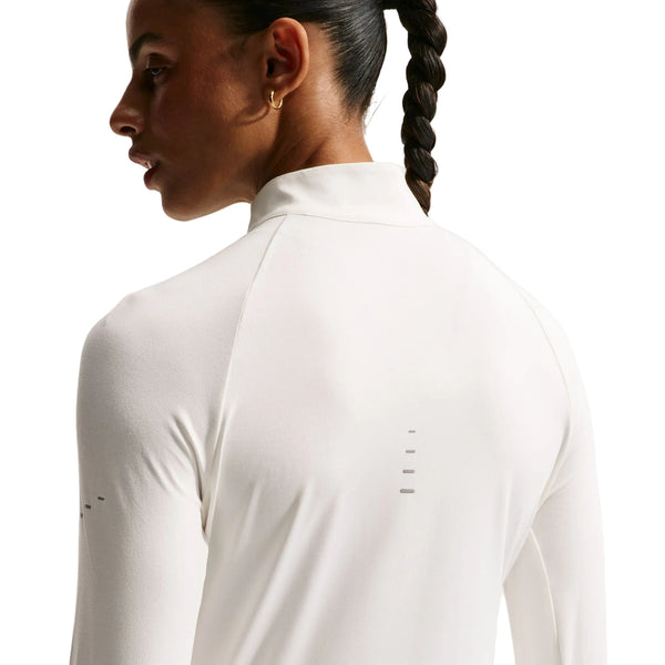 En person med en flettet hestehale ses set bagfra iført Nike Swift UV Half Zip-løbetrøje til kvinder i farven Sail/Reflective Silver. Den hvide, langærmede trøje har en høj krave og diskrete reflekterende detaljer. Billedet fremhæver pasformen og teksturen i det svedtransporterende stof, der er udviklet til at give fremragende komfort og UV-beskyttelse under udendørsaktiviteter.
