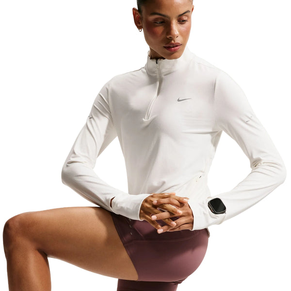 En person har en langærmet Nike Swift UV Half Zip-løbetrøje til kvinder i farven Sail/Reflective Silver på, der er fremstillet af svedtransporterende materiale og har UV-beskyttelse, kombineret med mauvefarvede shorts. Personen knæler med hænderne foldet og har et smartwatch på venstre håndled. Baggrunden er hvid.