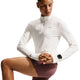 En person har en langærmet Nike Swift UV Half Zip-løbetrøje til kvinder i farven Sail/Reflective Silver på, der er fremstillet af svedtransporterende materiale og har UV-beskyttelse, kombineret med mauvefarvede shorts. Personen knæler med hænderne foldet og har et smartwatch på venstre håndled. Baggrunden er hvid.