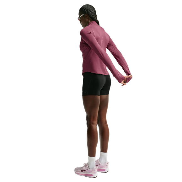 En person iført en Nike Swift UV Half Zip-løbetrøje til kvinder i farven Pink Smoke/Reflective Silver, sorte shorts, hvide sokker og lyserøde sneakers står og strækker armene bag ryggen foran en hvid baggrund.