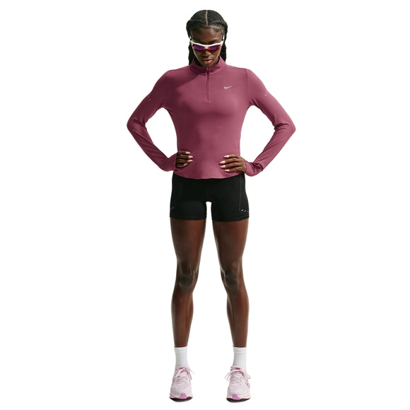 En person står med ansigtet vendt fremad og er iført en lyserød Nike Swift UV Half Zip-løbetrøje med lange ærmer (Dame, Pink Smoke/Reflective Silver) med UV-beskyttelse, sorte shorts, hvide sneakers og solbriller. Hænderne er lagt på hofterne, og håret er flettet. Baggrunden er ensfarvet hvid.
