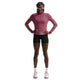 En person står med ansigtet vendt fremad og er iført en lyserød Nike Swift UV Half Zip-løbetrøje med lange ærmer (Dame, Pink Smoke/Reflective Silver) med UV-beskyttelse, sorte shorts, hvide sneakers og solbriller. Hænderne er lagt på hofterne, og håret er flettet. Baggrunden er ensfarvet hvid.