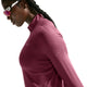 En person med flettet mørkt hår står i profil foran en hvid baggrund iført Nike Swift UV Half Zip-løbetrøje til kvinder i farven Pink Smoke/Reflective Silver. Personen bærer desuden reflekterende sportsolbriller.