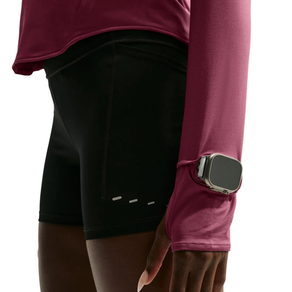 Der vises et billede af en person set fra underkroppen og armen, klædt i sorte shorts og en Nike Swift UV Half Zip-løbetrøje til kvinder i farven Pink Smoke/Reflective Silver, der har UV-beskyttelse. På håndleddet, over ærmet, bæres et smartwatch, og billedet er taget foran en hvid baggrund.