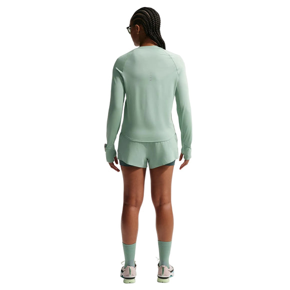 En dame med flettet hår, der vender væk fra kameraet, er iført en Nike Swift UV Long Sleeve Shirt (Dame, Steam/Reflective Silv) sammen med matchende shorts og sneakers på en almindelig hvid baggrund.
