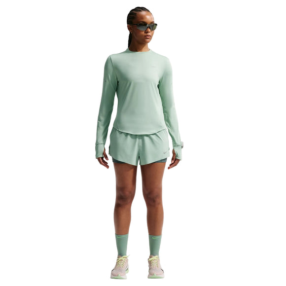 En kvinde med flettet hår og reflekterende sølvfarvede solbriller poserer selvsikkert i en Nike Swift UV langærmet skjorte til kvinder (Steam/Reflective Silver) på en almindelig hvid baggrund. Hun sætter den sammen med matchende shorts, høje sokker og sneakers.