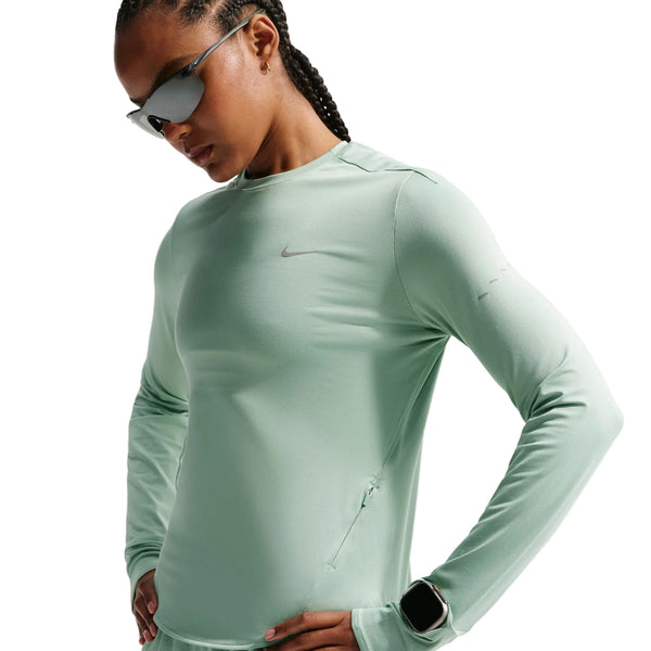En kvinde i Nikes Swift UV Long Sleeve Shirt til kvinder, i Steam/Reflective Silver, med lynlåslomme og spejlsolbriller står med hænderne på hofterne mod en ensfarvet baggrund.