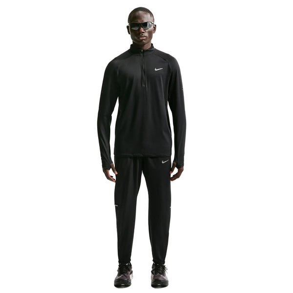 En person står med ansigtet fremad, iført Nike Stride Half Zip Midlayer-løbetrøje til herre i farven Black/Reflective Silv og matchende bukser, suppleret med sorte sko og mørke solbriller. Trøjen er perfekt til herretræning, og Nike-logoet er synligt både på trøjen og bukserne.
