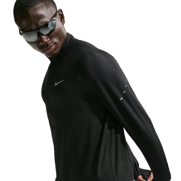 En person med mørke solbriller har en Nike Stride Half Zip Midlayer løbetrøje til mænd på, i farven sort/reflekterende sølv, med diskrete reflekterende detaljer. Stående foran en hvid baggrund og let vendt væk fra kameraet viser vedkommende, hvordan denne stilfulde og højtydende løbetrøje er perfekt til mænds træning.