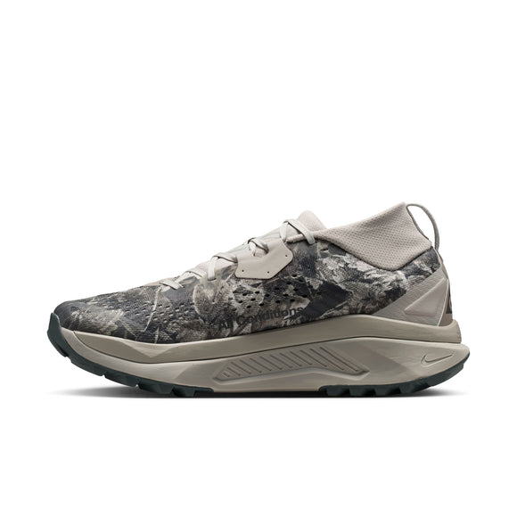 Nike ACG Zegama til mænd, vist i profil på en hvid baggrund, er en lavskåret sportssko inspireret af trail-løbesko-kategorien. Den kommer i farvekombinationen Spruce Fog/Black Spruce-Sea Glass-Black med et gråt og beige camouflagemønster, en tyk beige sål udstyret med Vibram Megagrip for overlegen greb, sort slidbane og snørelukning.