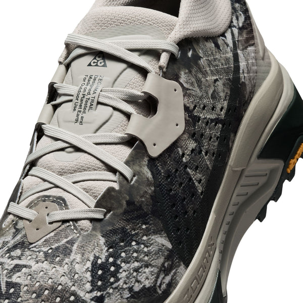 Nærbillede af Nike ACG Zegama trail-løbesko til mænd i farvekombinationen Spruce Fog/Black Spruce-Sea Glass-Black, med en overdel i gråt og sort mønstret mesh, hvide snørebånd og en synlig tunge med mærkat. Den lysegrå mellemsål kombineres med en ydersål med et lille orange indsnit og en robust Vibram Megagrip-sål, der sikrer forbedret greb – ideel til dem, der søger pålidelig ydeevne til trail-løb.