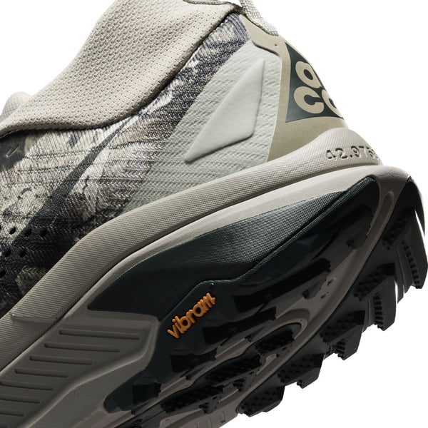 Nærbillede af hælen og ydersålen på Nike ACG Zegama til mænd i farvekombinationen Spruce Fog/Black Spruce-Sea Glass-Black, med et struktureret mønster, der giver godt greb, samt det sorte Vibram Megagrip-logo, stof med camouflagemønster og delvist Nike-logo i beige. Skoen fremstår slidstærk og designet til udendørs brug med et robust design.