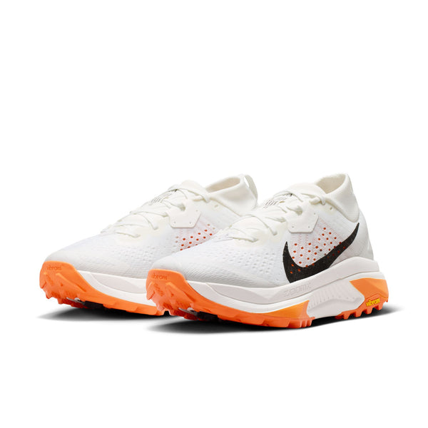 Et par Nike ACG Zegama trail-løbesko til damer i Summit White/Black-Phantom-Safety Orange, med sorte swoosh-logoer og orange Vibram Megagrip-såler, præsenteret mod en hvid baggrund.