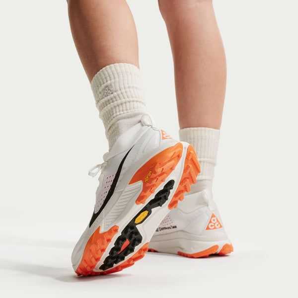 En person vises fra læggene og ned, iført hvide sokker og Nike ACG Zegama Dame-sko i Summit White/Black-Phantom-Safety Orange med orange og sorte såler, stående på en hvid flade.