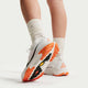 En person vises fra læggene og ned, iført hvide sokker og Nike ACG Zegama Dame-sko i Summit White/Black-Phantom-Safety Orange med orange og sorte såler, stående på en hvid flade.