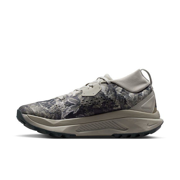 Et billede set fra siden af Nike ACG Zegama Dame trail-løbeskoen i farvekombinationen Spruce Fog/Black Spruce-Sea Glass-Black, med et camouflageagtigt blomstermønster, en struktureret Vibram Megagrip-sål, snørelukning og en tyk, polstret mellemsål. Nike-logoet er placeret tæt på hælen.