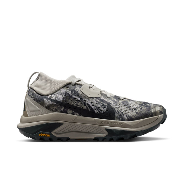 En Nike ACG Zegama trail-løbesko til kvinder i farvekombinationen Spruce Fog/Black Spruce-Sea Glass-Black, med en mønstret overdel i grå og sorte nuancer, en tyk sål, Vibram-logo på ydersålen med Vibram Megagrip-teknologi samt teksten »Zegama« på siden. Skoen vises i profil mod en hvid baggrund.