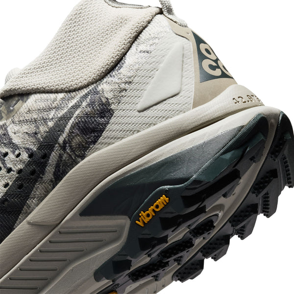 Nærbillede af hælen og sålen på en lys trail-løbesko, nærmere bestemt Nike Acg Zegama Dame i farvekombinationen Spruce Fog/Black Spruce-Sea Glass-Black, der viser et Vibram Megagrip-logo på det sorte slidbane og et delvist billede af skoens overdel med dens strukturerede designelementer.