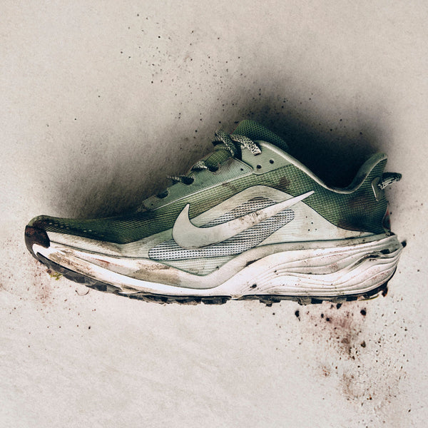 En Nike ACG Pegasus Trail Dame i Jade Horizon/Phantom-Light Silver ligger på siden mod en let baggrund, hvor snavs og mudder fremhæver dens robuste ReactX-skumdesign til det hårdeste terræn.