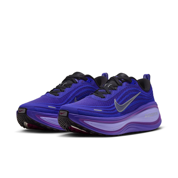 Et par Nike Vomero Plus Dame-løbesko i farvekombinationen BLACK/METALLIC SILVER-PERSIAN VIOLET har sorte snørebånd, Swoosh-logoer i metallisk sølv og en tyk mellemsål med farveovergang, der sikrer maksimal komfort. Skoene er afbilledet vinklet mod venstre på en hvid baggrund.