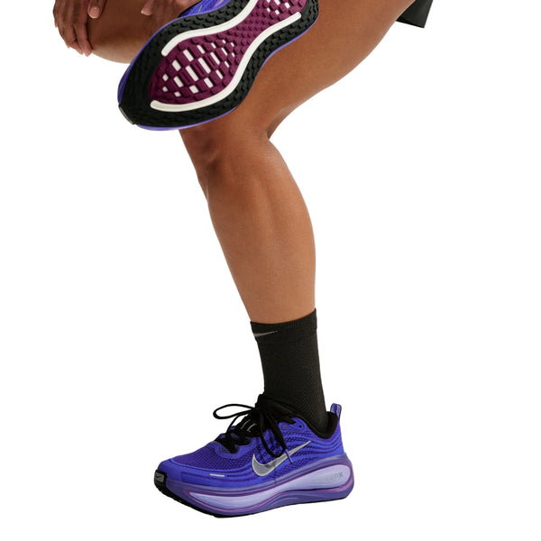En person iført sorte strømper og Nike Vomero Plus Dame-løbesko i farvekombinationen BLACK/METALLIC SILVER-PERSIAN VIOLET, med et synligt Nike-logo, løfter det ene ben og afslører den strukturerede sål, der er designet til maksimal komfort. Kun den nederste halvdel af personens krop ses på en hvid baggrund.