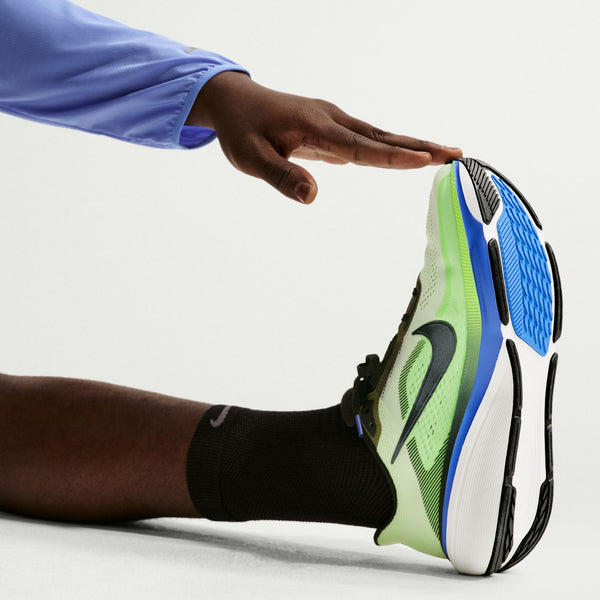 En person i sort strømpe og Nike Pegasus 42 Herre, Volt Tint/Black Spruce-Sapphire med ReactX-skummellemsål strækker sig og rører ved toppen af sin sko, armen er klædt i et lyseblåt ærme.