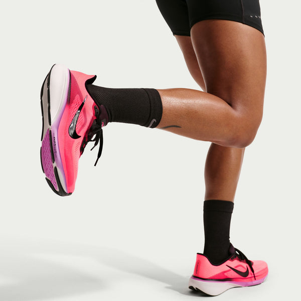 En person iført sorte shorts, sorte sokker og Nike Pegasus 42 Dame løbesko i Lava Glow/Black-Flash Crimson-Lt Magenta står med det ene ben bøjet bagover, som om han strækker sig eller forbereder sig på at løbe.