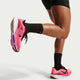En person iført sorte shorts, sorte sokker og Nike Pegasus 42 Dame løbesko i Lava Glow/Black-Flash Crimson-Lt Magenta står med det ene ben bøjet bagover, som om han strækker sig eller forbereder sig på at løbe.