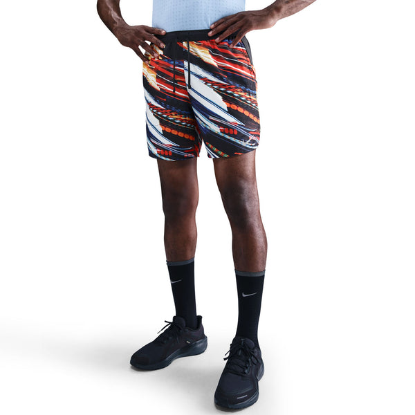En person står med hænderne på hofterne iført en lyseblå skjorte, Nike Nk Df Stride 7In Bf Shrt Op2 shorts i White/Multi Color/Reflective Silver, sorte knæhøje Nike-sokker og sorte sportssko på en hvid baggrund.