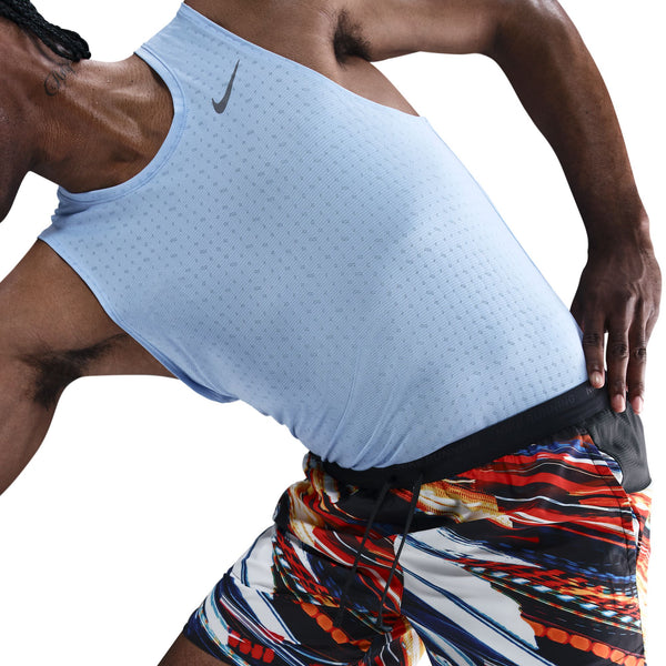 En person iført en lyseblå Nike-tanktop og Nike Nk Df Stride 7In Bf Shrt Op2-shorts i White/Multi Color/Reflective Silver strækker sig med den ene hånd på hoften og viser overkroppen og en del af benene mod en hvid baggrund.