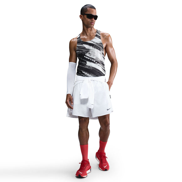 En mand står og kigger fremad iført en Nike M Nk Df Fast Singlet til mænd i Photon Dust/Reflective Silver, parret med hvide sportsshorts, røde sokker, røde sneakers, et hvidt ærme på højre arm, sorte solbriller og en hvid sweatshirt bundet i taljen.