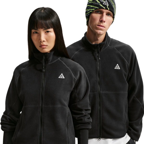 To personer står side om side, begge iført Nike ACG »Wolf Tree«-jakken med gennemgående lynlås til mænd i farvekombinationen Sort/Sort/Sort/Summit White, der er fremstillet af Polartec-fleece og har et diskret trekantet logo på brystet. Personen til højre bærer desuden en mønstret hue. Begge ser mod kameraet med neutrale ansigtsudtryk og fremviser dermed disse alsidige Nike-fleecejakker til mænd.