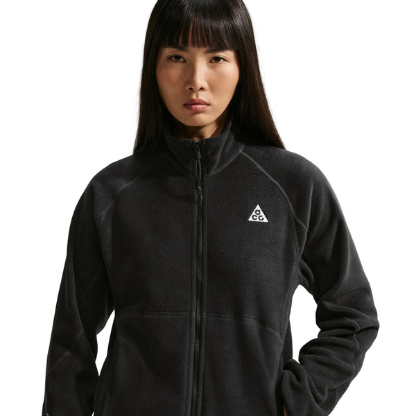 En person med langt, mørkt hår har Nike ACG »Wolf Tree«-jakken med gennemgående lynlås til mænd i farvekombinationen Sort/Sort/Sort/Summit White på. Jakken er fremstillet af blødt Polartec-fleece og har et lille trekantet logo på venstre bryst på en ensfarvet hvid baggrund.