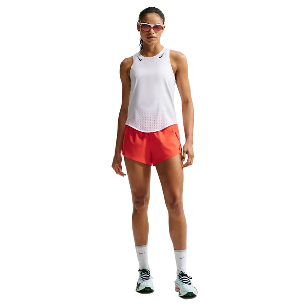 En person ser frem for sig iført en hvid/sort Nike AeroSwift Running Singlet (Dame), lyse orange shorts, hvide sokker, hvide løbesko med grønne detaljer og solbriller.