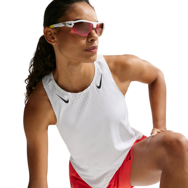 En person iført en hvid/sort Nike AeroSwift Running Singlet til kvinder poserer med et løftet knæ, sammen med røde shorts og reflekterende sportssolbriller mod en hvid baggrund.