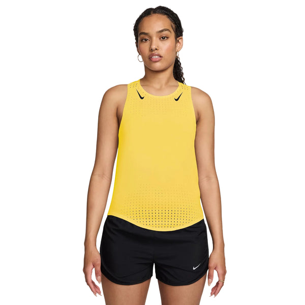 En kvinde står og kigger fremad iført en Nike AeroSwift Running Singlet, Dame, i Citron Pulse/Black med Dri-FIT ADV-teknologi og sorte Nike-shorts, begge med mærkets logo, på en almindelig hvid baggrund.