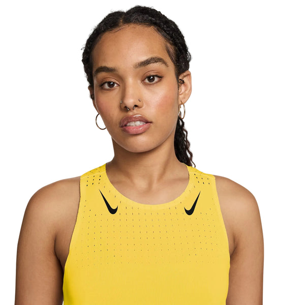 En person med medium hud, flettet hår og øreringe bærer Nike AeroSwift Running Singlet til damer i Citron Pulse/Black. Dri-FIT ADV-teknologi og sorte swoosh-logoer skiller sig ud mod en almindelig hvid baggrund.