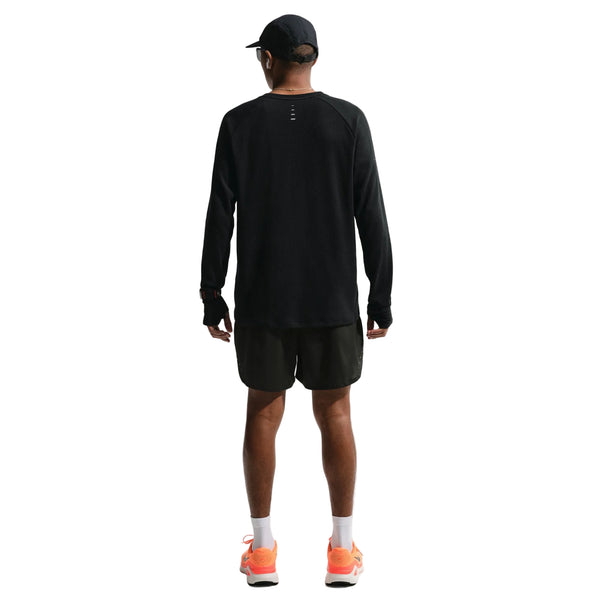 En person står med front mod kameraet iført en sort langærmet skjorte, en sort kasket, hvide sokker, orange løbesko og sorte Nike Stride Plus 5" brief-lined løbeshorts til mænd på en almindelig hvid baggrund.