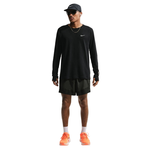 En mand ser frem for sig i en sort langærmet Nike-trøje, sort Nike-kasket og Stride Plus 5" Brief-Lined Running Shorts (Herre, Black) fra Nike. Han har orange sneakers, solbriller og en tynd halskæde på mod en hvid baggrund.