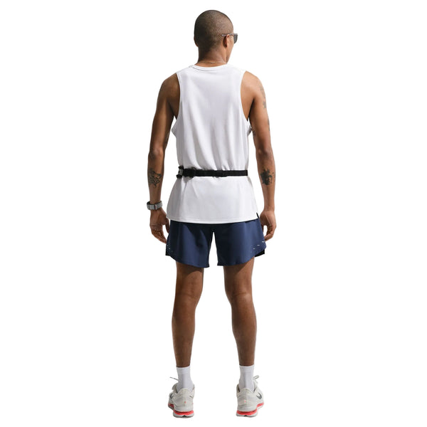 En person står med ryggen til og er iført en hvid ærmeløs skjorte, Nike Stride 2In1 7" løbeshorts til mænd i Midnight Navy/Reflective Silver, hvide sokker og sneakers, en sort bæltetaske, et ur og solbriller på en almindelig hvid baggrund.