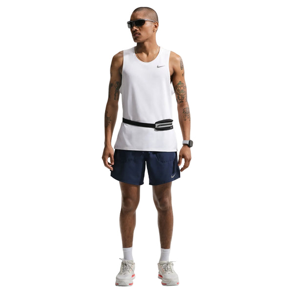 En mand med solbriller og en hvid ærmeløs top bærer Nike Stride 2In1 7" løbeshorts til mænd i Midnight Navy/Reflective Silver, plus hvide sokker, sportssko, tatoveringer, et smartur og et sort taljebælte på en hvid baggrund.