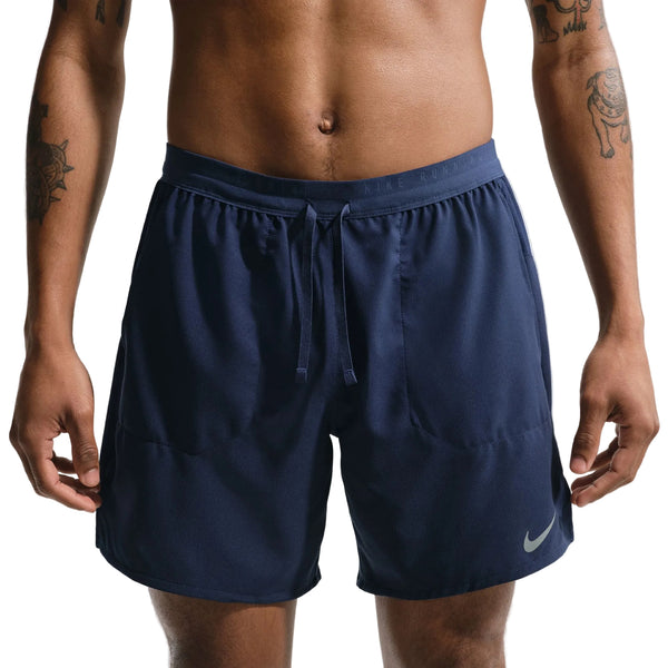 En person iført Nike Stride 2In1 7" løbeshorts til herrer i Midnight Navy/Reflective Silver, med elastisk linning, snøre og Nike-logo på venstre ben. Overkroppen og de tatoverede arme er delvist synlige.