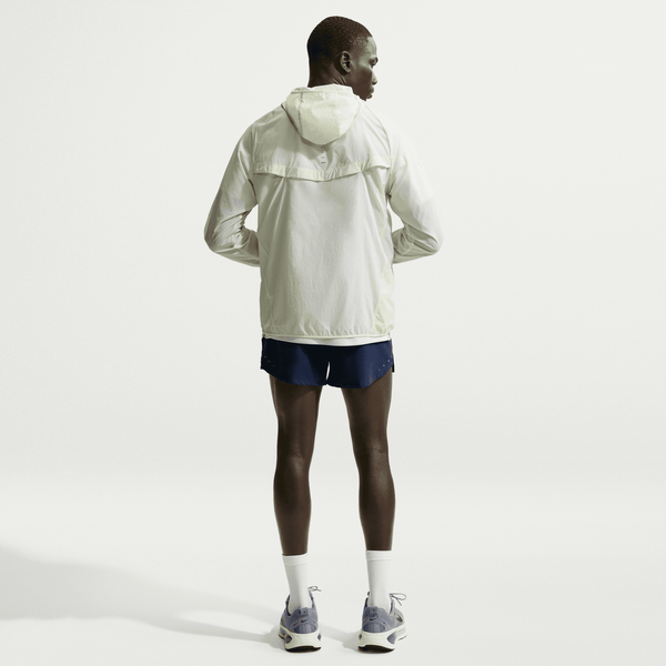 En person står med ryggen til kameraet iført en lys jakke med hætte, Nike Stride 5" Brief-Lined Running Shorts til mænd i Midnight Navy/Reflective Silver, hvide sokker og grå sportssko på en almindelig hvid baggrund.