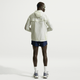 En person står med ryggen til kameraet iført en lys jakke med hætte, Nike Stride 5" Brief-Lined Running Shorts til mænd i Midnight Navy/Reflective Silver, hvide sokker og grå sportssko på en almindelig hvid baggrund.