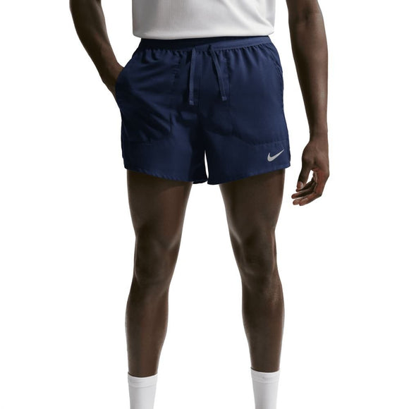 En person står på en almindelig baggrund med hænderne i lommerne og viser kun sin underkrop og sine ben i Nike Stride 5" Brief-Lined Running Shorts til mænd, Midnight Navy/Reflective Silver.