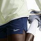 En person i lys jakke og Nike Stride 5" Brief-Lined Running Shorts, Herre, Midnight Navy/Reflective Silv strækker benet, mens han holder en grå og hvid sneaker. Kun den nederste halvdel af kroppen er vist mod en almindelig baggrund.