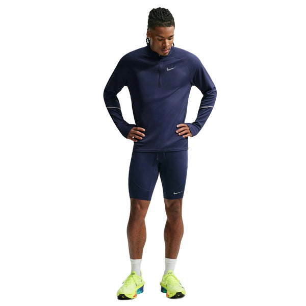 En person står med ansigtet vendt fremad og hænderne på hofterne. Personen er iført en marineblå Nike-trøje med lange ærmer samt Nike Stride Running Half Tights til mænd i farvekombinationen Midnight Navy/Reflective Silver, suppleret med hvide sokker og neongule løbesko. Baggrunden er ensfarvet hvid.