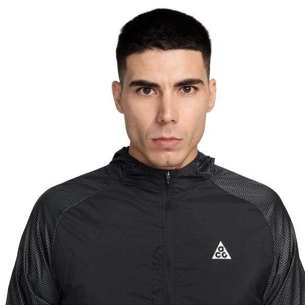 En mand med kort mørkt hår er iført en sort Nike ACG Uv Aireez Jkt (Black/Black/Summit White) med et hvidt trekantet ACG-logo på brystet, der står på en almindelig hvid baggrund.