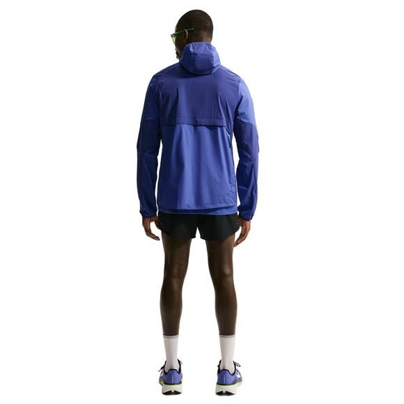 En person står med ryggen til i en Nike Stride Plus løbejakke, Herre, Deep Royal/Lapis/Reflective Silver, sammen med sorte shorts, hvide sokker og løbesko. Baggrunden er almindelig hvid.